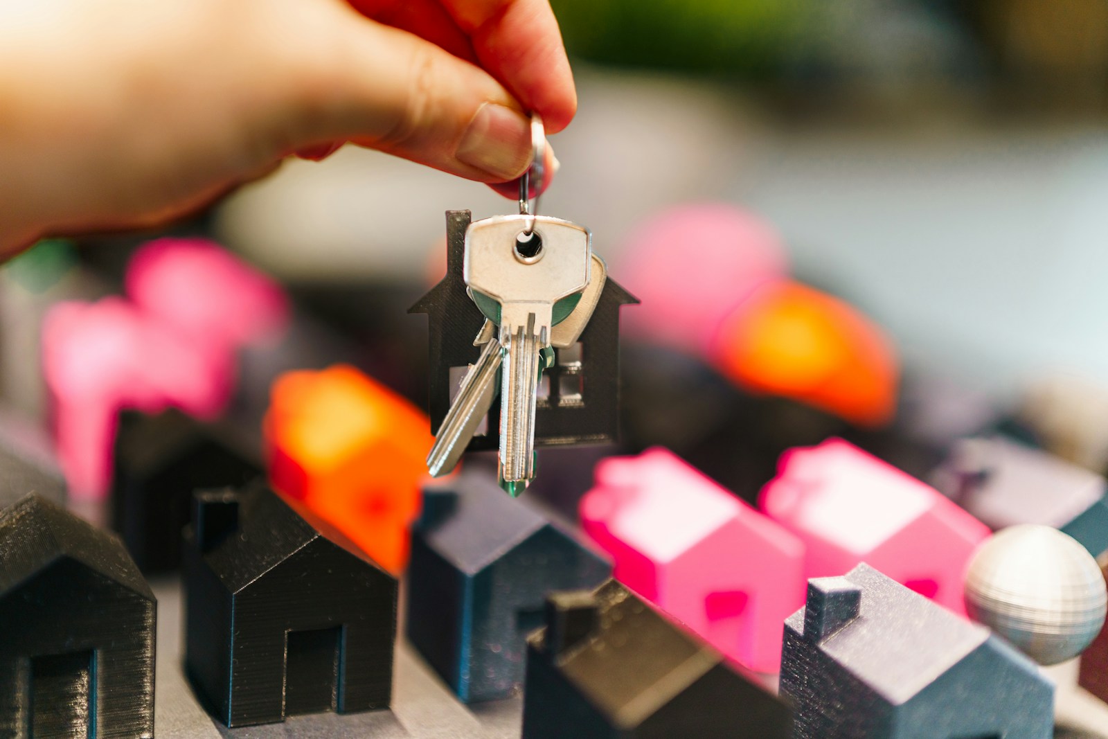 Hand holding silver house keys above colorful pink and orange model homes