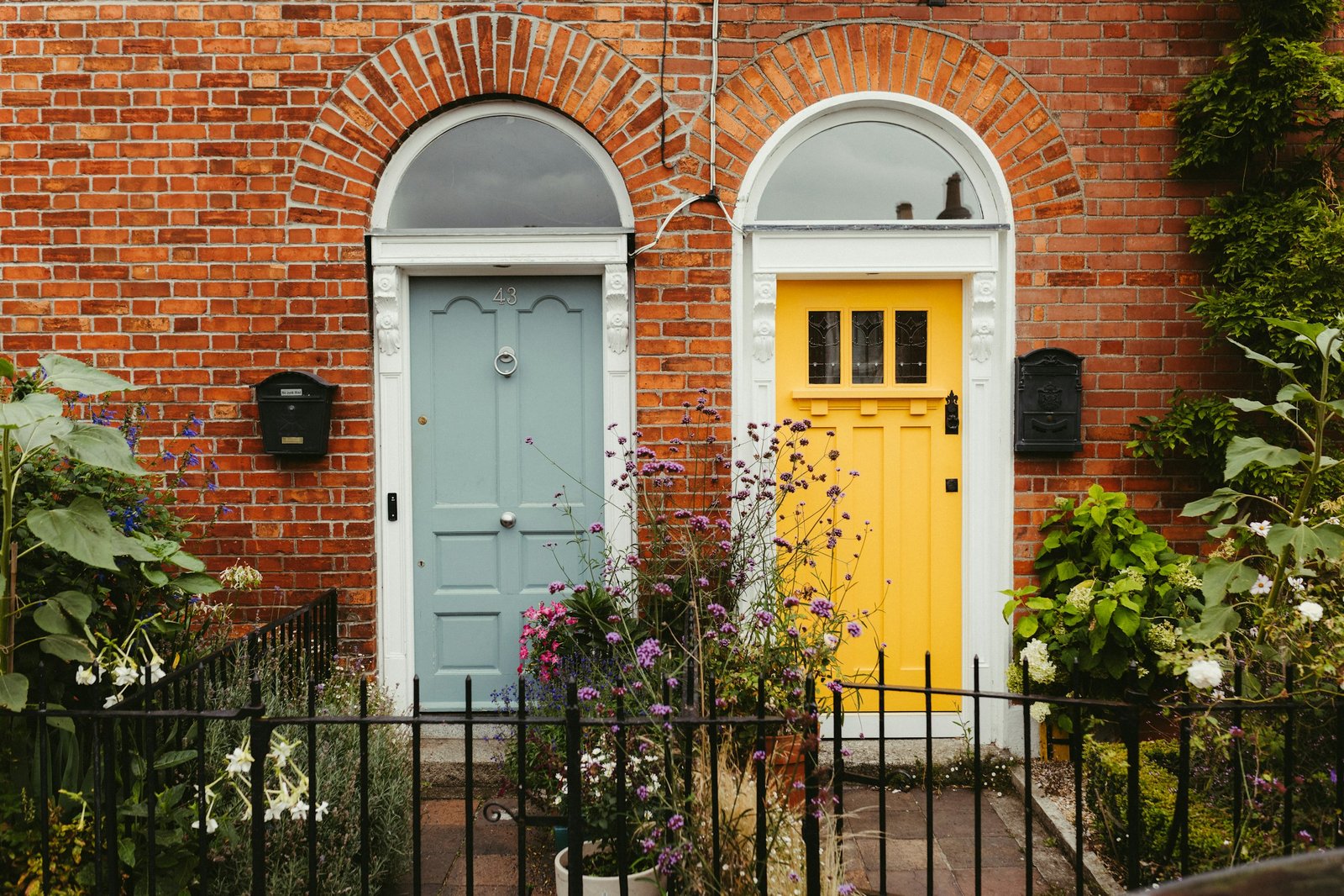 A bright yellow front door and a pale blue front door on a brick house with flowers along the railing