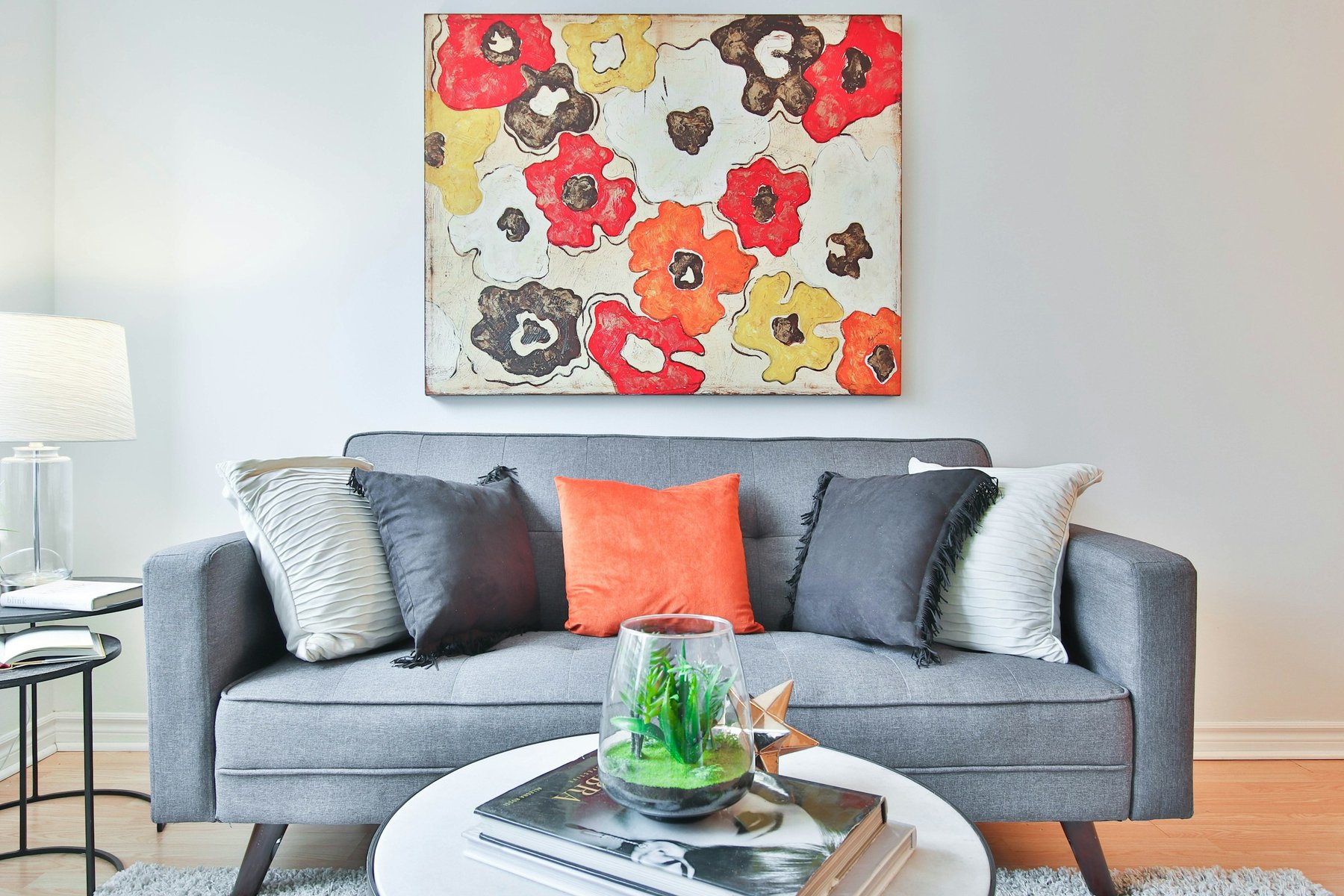 A bright living room with a gray sofa, orange and yellow pillows and a large floral painting