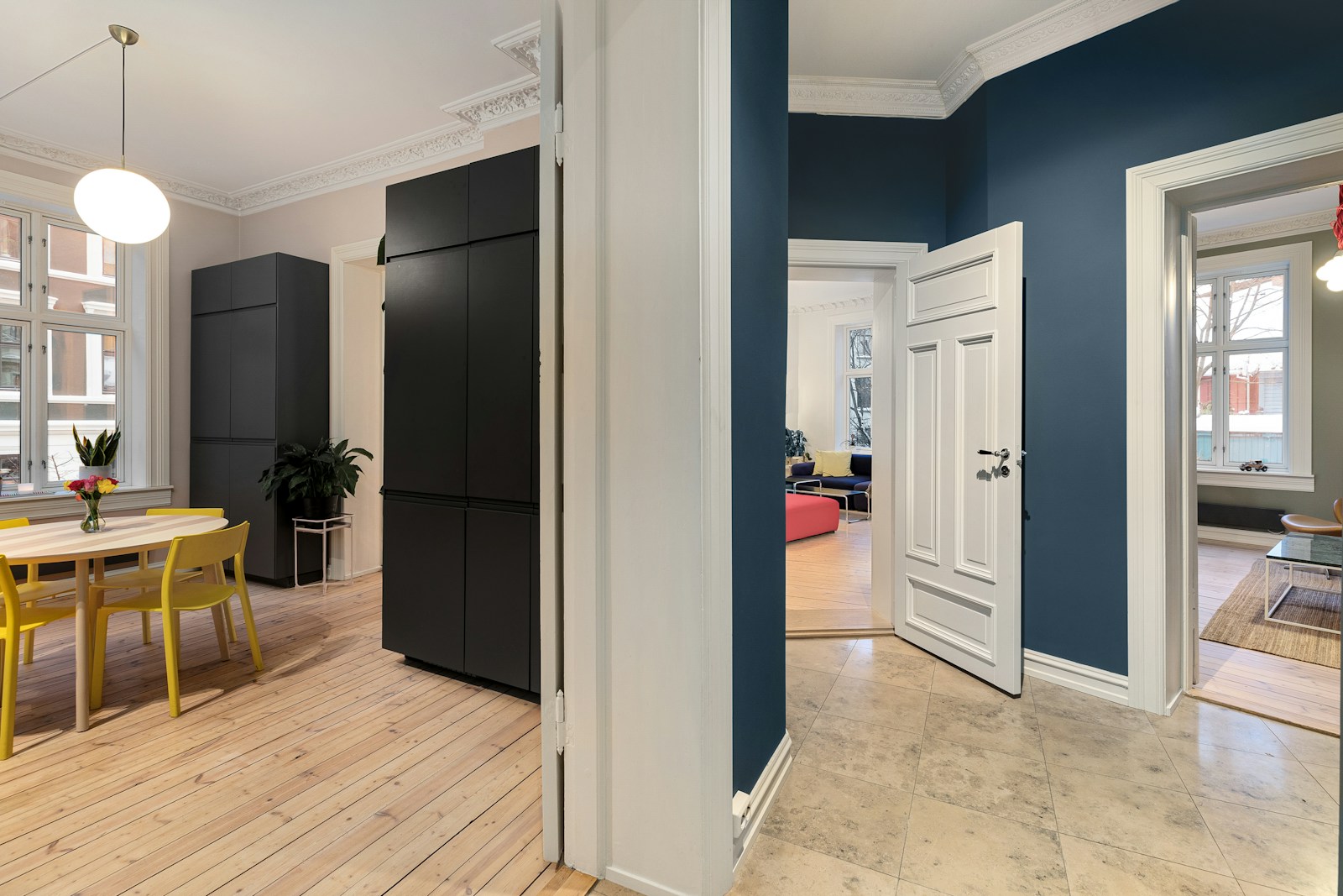 A tidy navy-blue hallway with white trim beside a bright dining area with yellow chairs