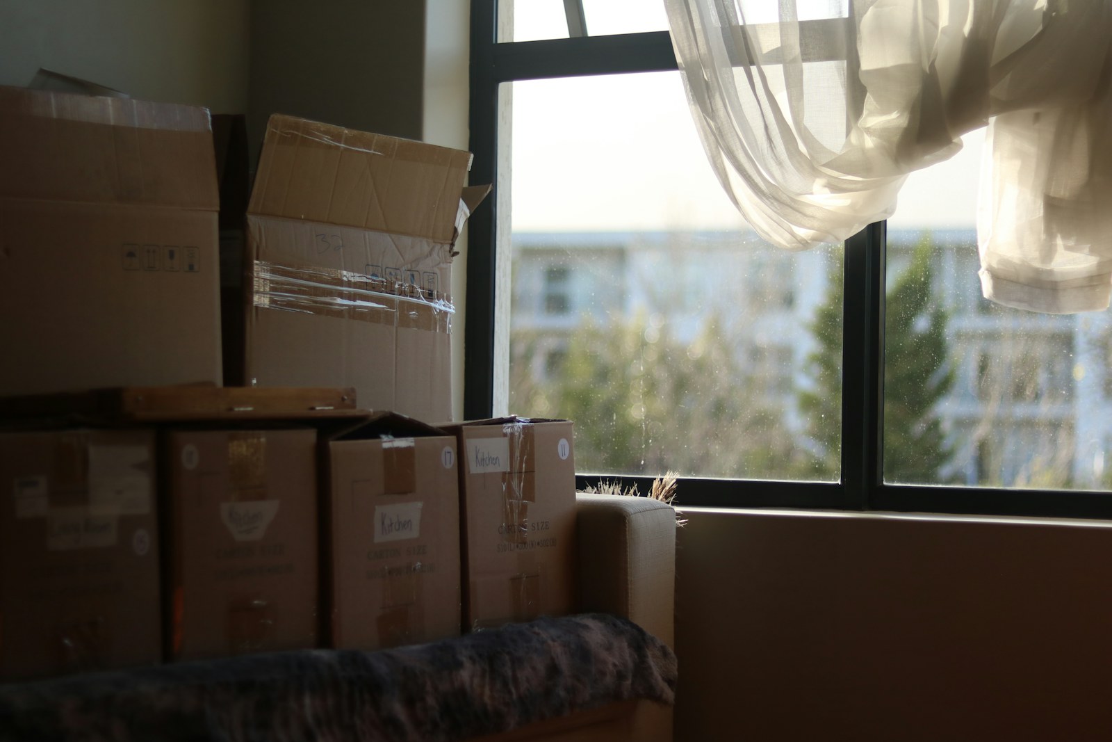 Stacked cardboard moving boxes beside a sunny window with a sheer curtain