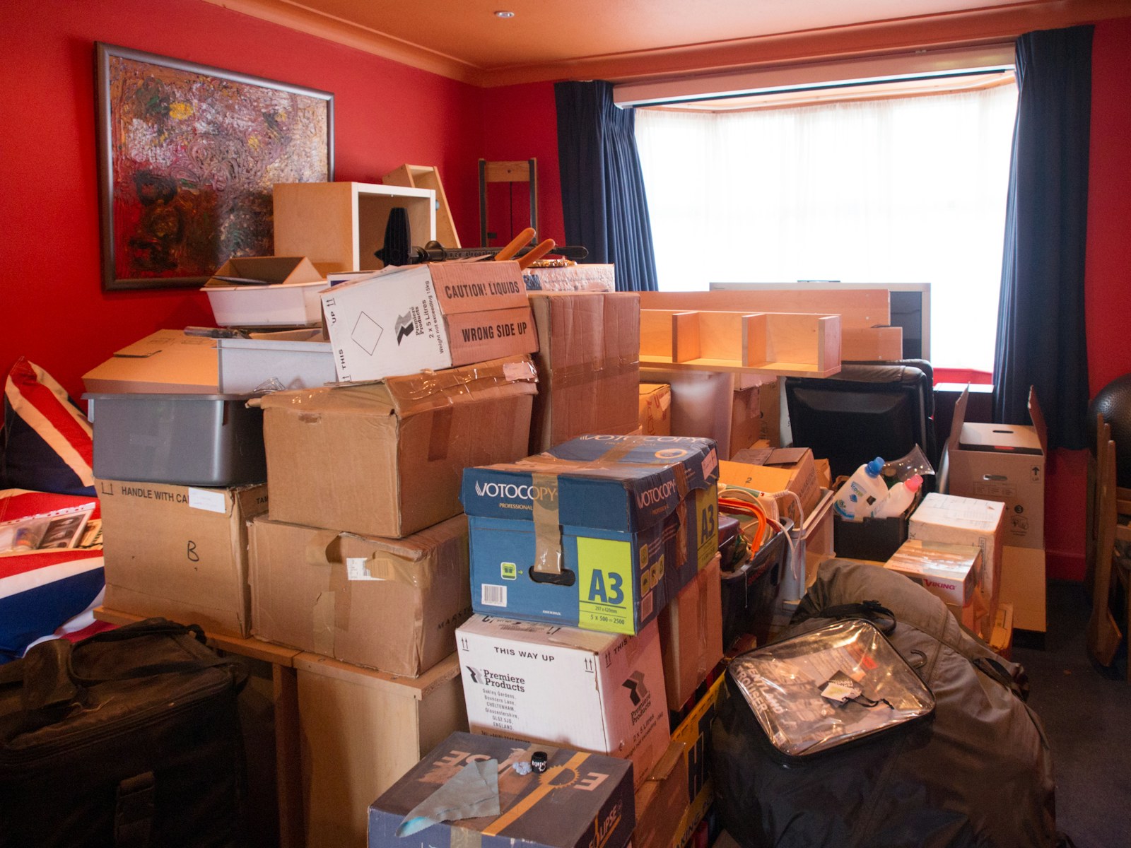 Moving boxes and bags piled in a living room with red walls and a bay window