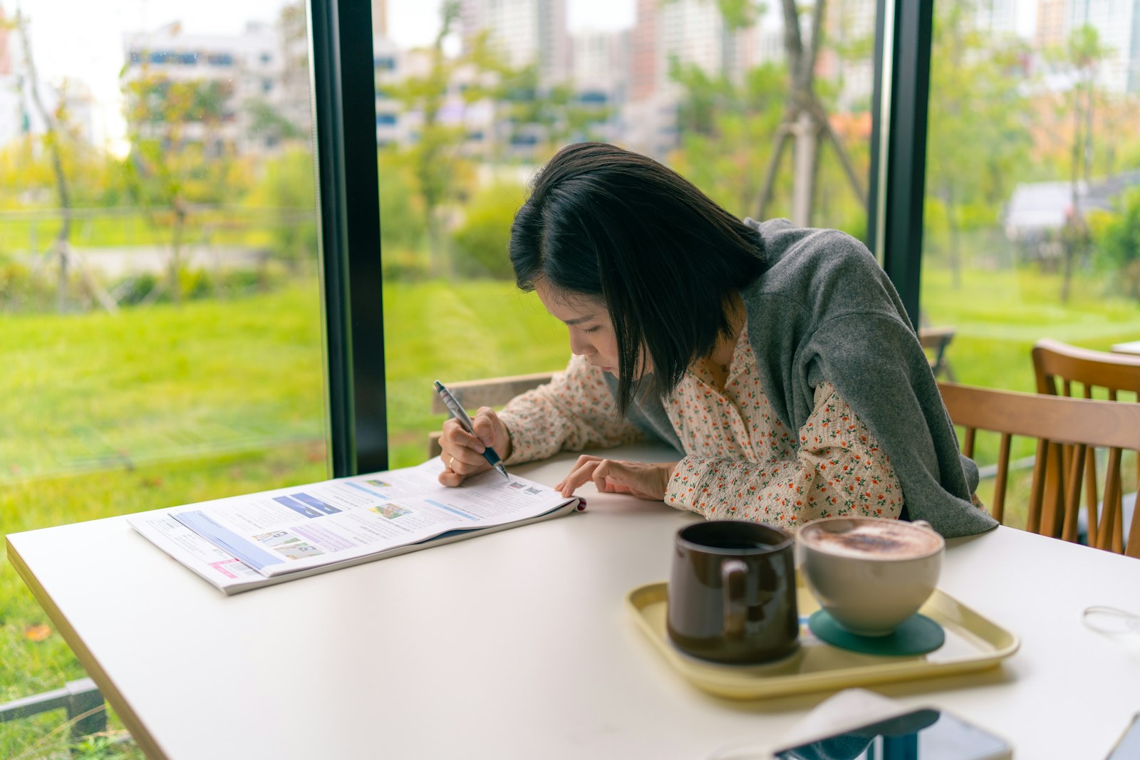 Woman writing at a table by a bright window with greenery outside